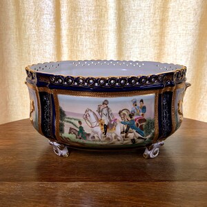 May include: A large, blue and gold porcelain bowl with a decorative scene of people on horseback. The bowl has a scalloped edge and intricate details.