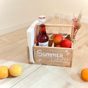 Może przedstawiać: Drewniany pojemnik z butelką czerwonego płynu, dwoma szklankami, serwetkami i morelami. Pojemnik ma napis "Summer Vibes" i grafikę palmy. Suche kwiaty są przymocowane do uchwytu.