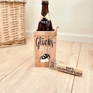 Może przedstawiać: Drewniany uchwyt na butelkę piwa z napisem "Glücks Pils" i grafiką grzyba. Butelka piwa znajduje się wewnątrz uchwytu. Obok znajduje się drewniany otwieracz do butelek z napisem "ken's Lieblingswerkzeug".