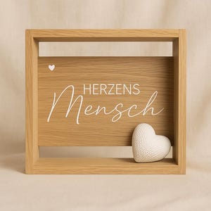 Może przedstawiać: Drewniany szyld w ramie z napisem "HERZENS Mensch" w białym skrypcie na jasnobrązowym tle. Małe białe serce znajduje się na górze, a teksturowane białe serce znajduje się po prawej stronie.