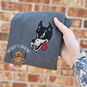 May include: A grey folded sweatshirt with an embroidered cartoon wolf head and a red tongue. The sweatshirt has the text "HENRY'S HOUSE EMBROIDERY" and a logo of a golden retriever dog surrounded by flowers.