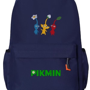 May include: A navy blue backpack featuring a graphic of three Pikmin characters in blue, yellow, and red colors. The front pocket displays the green text PIKMIN with a small white flower icon on the letter P. It has black shoulder straps and orange zipper pulls.