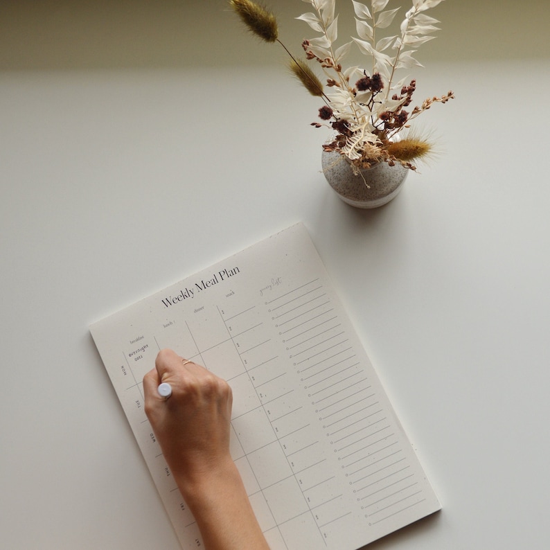 A female writing on her Weekly Meal Plan notepad. The notepad is on a desk with a vase of dried flowers. Each Weekly Meal Plan page has space for three meals and two snacks every day of the week and includes a dedicated space for your grocery list.