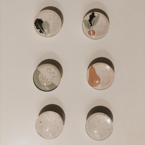 May include: Six round glass magnets with various designs. The magnets feature a green leaf, a turnip, a pear, a green and white design with the text "grow dear you", a white magnet with brown speckles, and another white magnet with brown speckles.