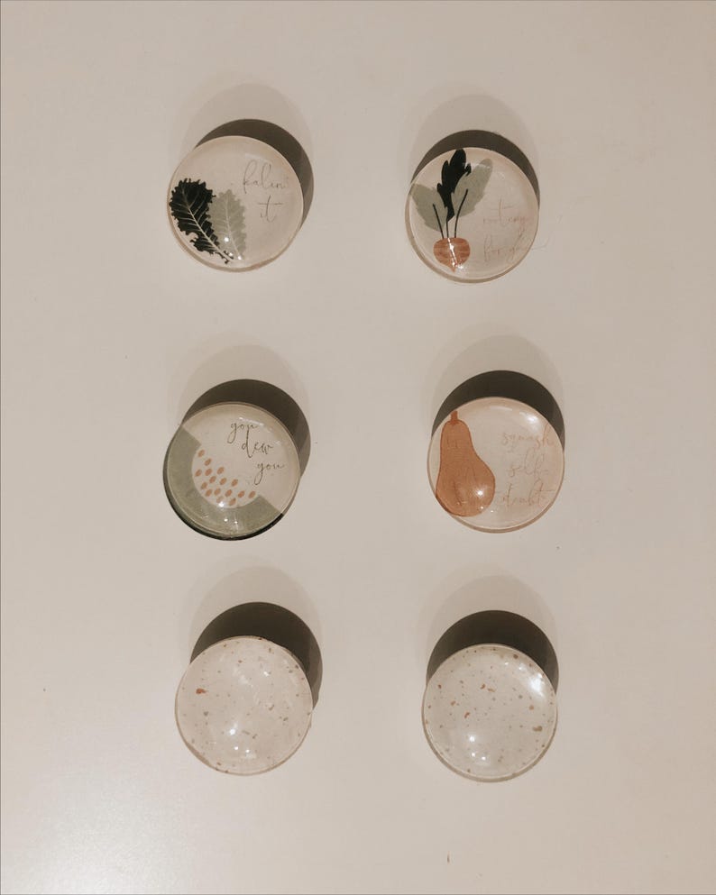 May include: Six round glass magnets with various designs. The magnets feature a green leaf, a turnip, a pear, a green and white design with the text "grow dear you", a white magnet with brown speckles, and another white magnet with brown speckles.