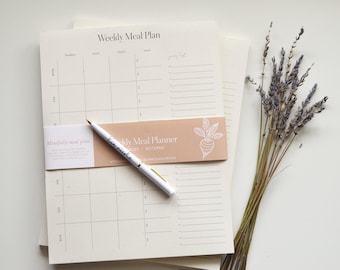 May include: A stack of weekly meal planner notepads with a white pen and a bunch of dried lavender flowers. The notepad has a light brown background with the text "Weekly Meal Planner" and a small illustration of a beet.