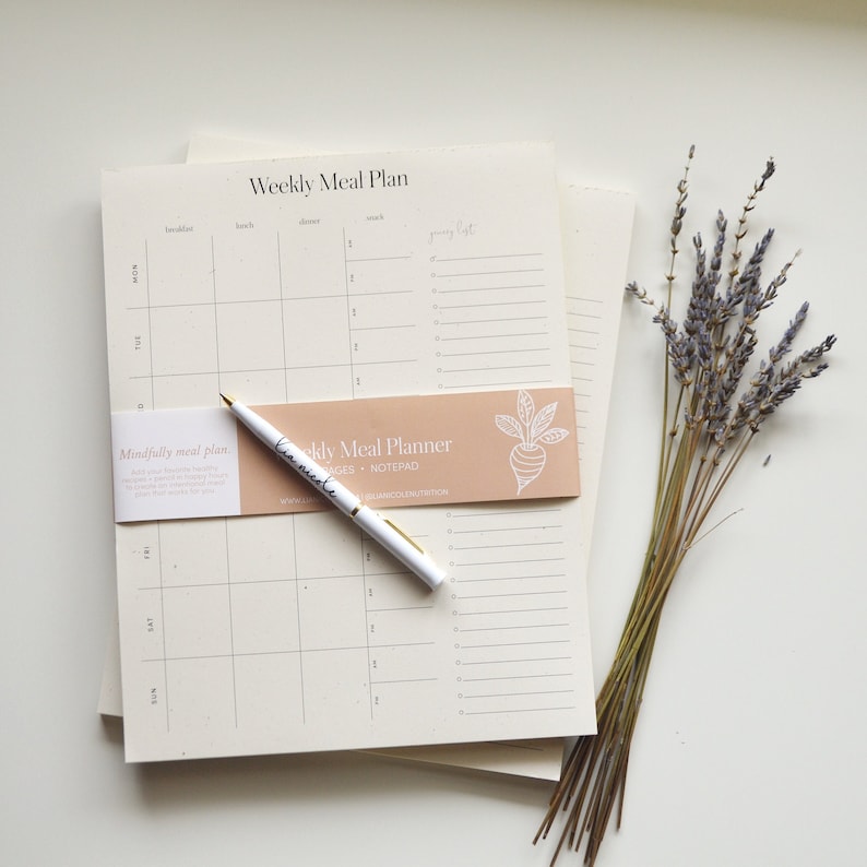 The Weekly Meal Plan notepad lying on a desk with a pen and lavender. Each Weekly Meal Plan page has space for three meals and two snacks every day of the week and includes a dedicated space for your grocery list.