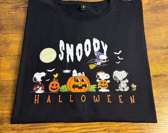 May include: A black t-shirt featuring a graphic print with the text SNOOPY HALLOWEEN. The design includes Snoopy dressed as a vampire, a witch on a broomstick, and a mummy, alongside Woodstock and several jack-o'-lanterns under a full moon.
