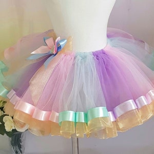 Può includere: Una gonna tutu di tulle arcobaleno con più strati di bordatura in nastro di raso. La gonna è esposta su un manichino bianco.