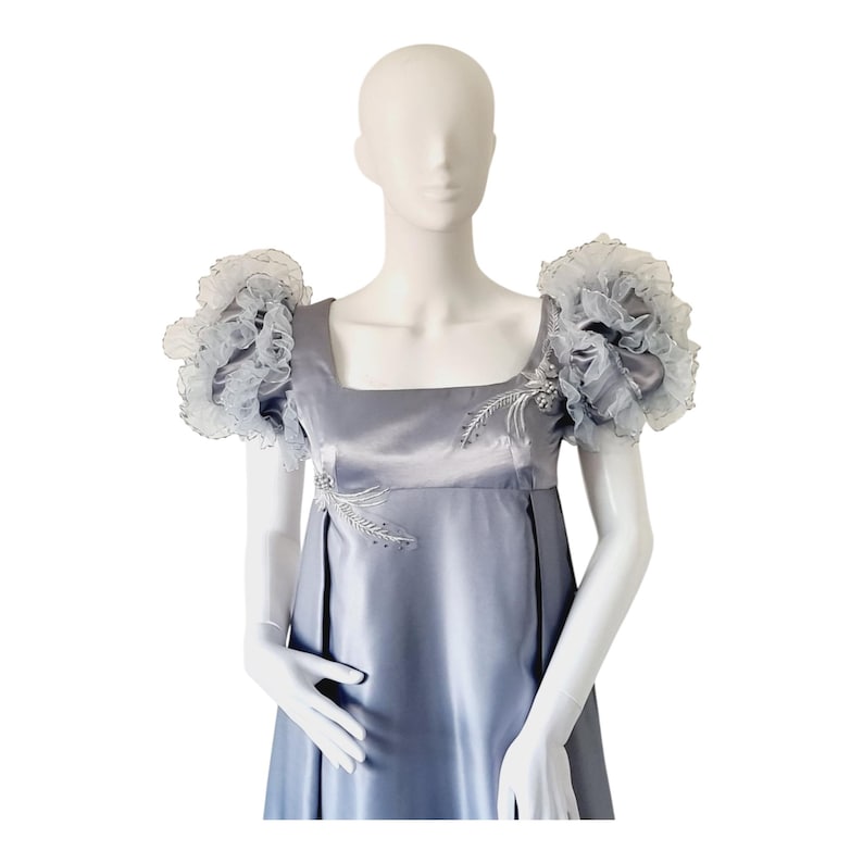 May include: A floor-length satin dress in a muted silver-blue hue, featuring a square neckline and voluminous, ruffled organza puff sleeves. The bodice is adorned with delicate silver embroidery and beadwork accents on the chest and shoulder.