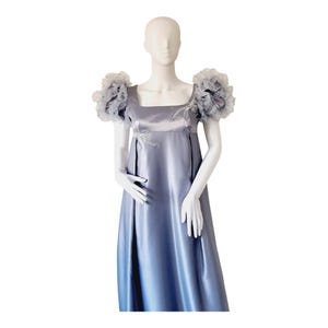May include: A floor-length periwinkle blue satin dress featuring a square neckline, empire waist, and voluminous ruffled cap sleeves. The bodice is accented with delicate silver-toned embroidery and beadwork detailing on the chest area.