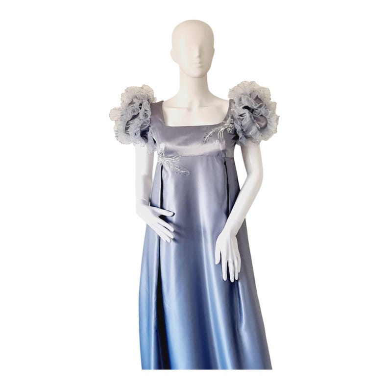 May include: A floor-length periwinkle blue satin dress featuring a square neckline, empire waist, and voluminous ruffled cap sleeves. The bodice is accented with delicate silver-toned embroidery and beadwork detailing on the chest area.