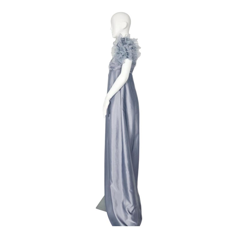 May include: A floor-length, slate blue sleeveless gown featuring a high waistline and a bodice adorned with layered, ruffled tulle detailing at the shoulders. The skirt is constructed from a smooth, satin-like fabric that falls in a straight line.