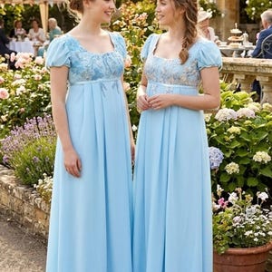 May include: Two floor-length light blue dresses featuring empire waistlines, short puff sleeves, and square necklines. The bodice of each dress is adorned with intricate silver and blue floral embroidery, while the skirt flows in a simple, pleated design.