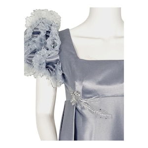 May include: A slate gray satin dress featuring a square neckline and a prominent, voluminous ruffled sleeve made of sheer organza. The bodice is accented with a delicate silver beaded floral appliqu&eacute; detail on the side, adding texture to the smooth fabric.
