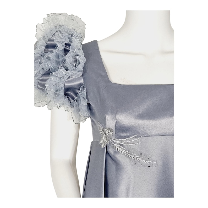 May include: A slate gray satin dress featuring a square neckline and a prominent, voluminous ruffled sleeve made of sheer organza. The bodice is accented with a delicate silver beaded floral appliqu&eacute; detail on the side, adding texture to the smooth fabric.