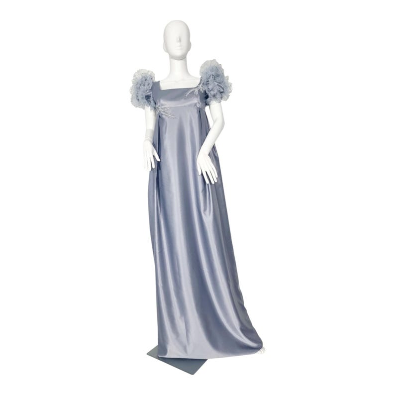 May include: A floor-length, slate blue satin gown featuring a square neckline and voluminous, ruffled tulle sleeves. The dress has a high-waisted empire silhouette with a smooth, flowing skirt that extends to the floor, displayed on a mannequin.