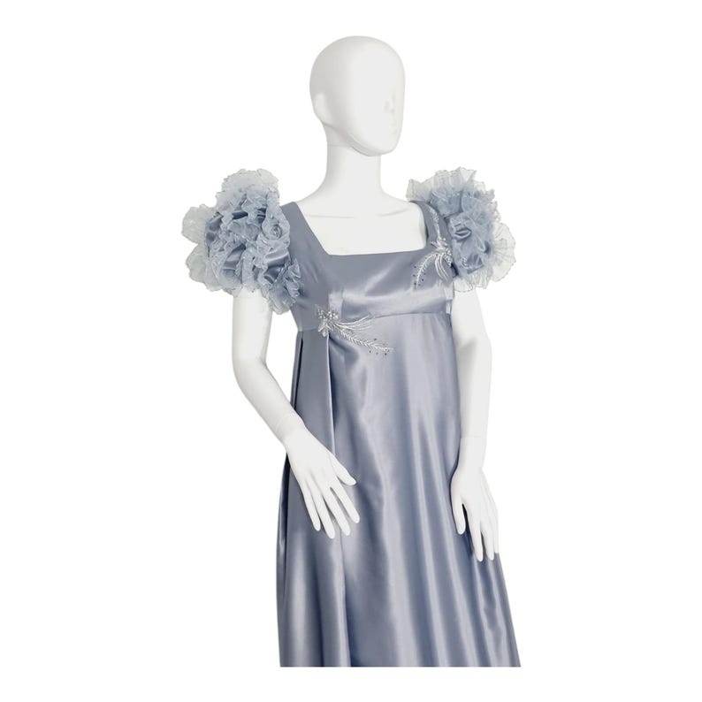May include: A floor-length, slate blue satin dress featuring a square neckline and an empire waist. The garment includes voluminous, ruffled tulle cap sleeves and delicate silver-toned embroidery accents on the bodice and shoulders.