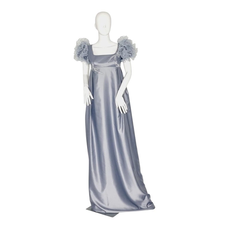 May include: A floor-length, periwinkle blue satin dress featuring a square neckline, an empire waistline, and voluminous, ruffled tulle cap sleeves. The bodice is accented with delicate, light-colored floral embroidery near the neckline.