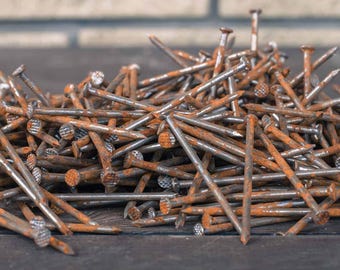 May include: A large pile of metal nails with visible rust and oxidation. Each nail features a flat, circular head and a pointed tip. The nails are scattered in a disorganised heap, showing varying degrees of corrosion across their surfaces.