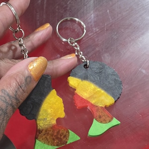May include: Two keychains featuring colorful profiles of a person. The keychains have a silver chain and ring. The profiles are painted with black, yellow, red, green, and brown colors. The background is red.