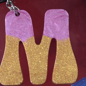 May include: A glittery letter "M" keychain. The top half is a pinkish-purple color, and the bottom half is gold. The keychain is attached to a silver chain. The background is a dark red color.