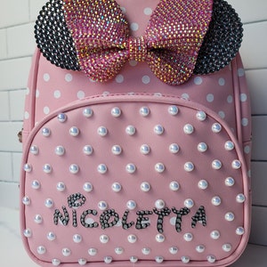 May include: Pink polka dot backpack with a large sparkly pink bow and black rhinestone ears. The backpack has a white pearl design and the name "Nicoletta" is spelled out in rhinestones.