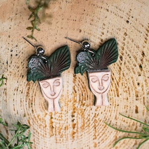 May include: A pair of ceramic earrings featuring a stylized face with green leafy details. The earrings are on a wooden surface.