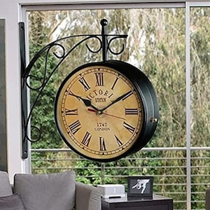 May include: A double-sided black metal clock with a cream-colored face, Roman numerals, and the text "VICTORIA STATION 1747 LONDON." The clock is mounted on a decorative bracket, suitable for indoor or outdoor use.