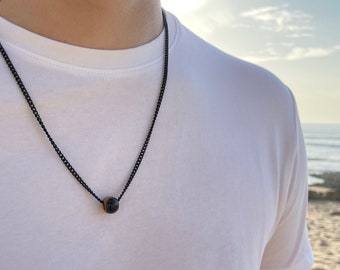 Puede incluir: Un collar de cadena negra con un solo colgante de cuentas redondas de color marrón oscuro. El collar se lleva sobre una camiseta blanca, con un fondo de playa y océano.