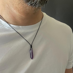 May include: A purple amethyst crystal pendant shaped like a pointed hexagonal prism, suspended from a thin black cord necklace. The crystal features a deep violet hue and a smooth, polished surface, hanging centered on a plain white t-shirt.