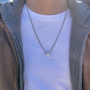 Puede incluir: Un collar de cadena plateada con un pequeño colgante rectangular. El colgante parece estar hecho de una piedra o material de color claro. La persona de la imagen lleva una camiseta blanca, una chaqueta con capucha gris y una chaqueta de cuero marrón.
