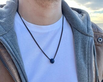 Puede incluir: Un collar que presenta una cadena de metal negro fina y un colgante de cuenta pequeña, lisa y de color azul oscuro. La cuenta tiene un acabado pulido y está centrada en la cadena, que descansa sobre una camiseta blanca debajo de una sudadera gris y una chaqueta marrón.