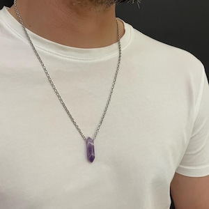 Pode incluir: Um colar de corrente prateada com um pingente de cristal de ametista roxa. O cristal é de cor roxa profunda e é cortado em forma hexagonal. O colar é usado sobre uma camiseta branca.