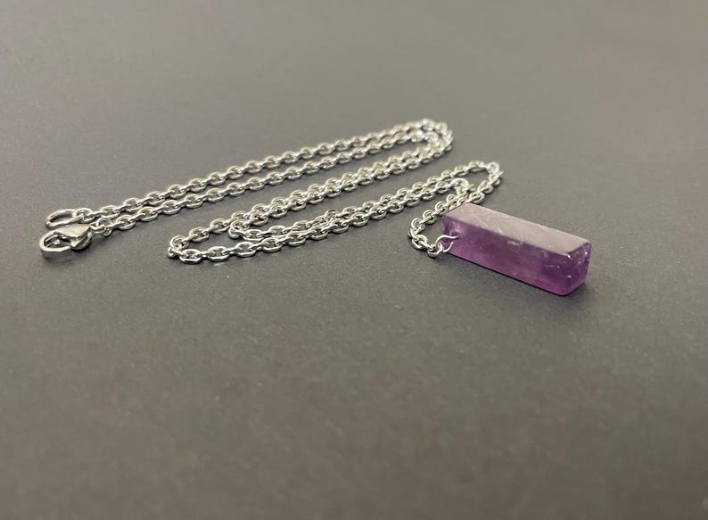 May include: A silver-toned metal chain necklace featuring a rectangular, polished purple amethyst gemstone pendant. The pendant is attached to the chain by a small metal loop, and the chain includes a lobster claw clasp at the end.
