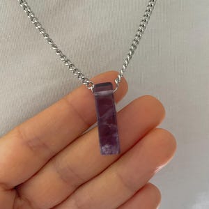 May include: A rectangular purple stone pendant with white marbling suspended from a silver-toned metal chain. The pendant features a smooth, polished finish and is attached to the chain by a small metal loop at the top of the stone.