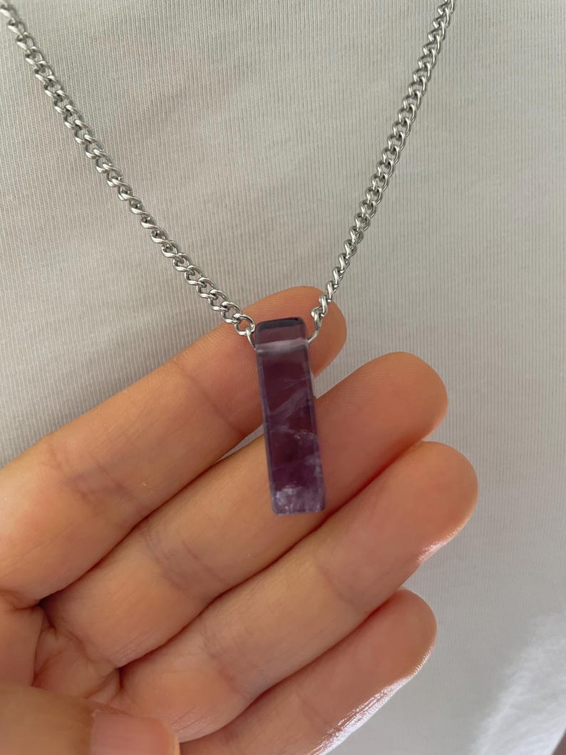May include: A rectangular purple stone pendant with white marbling suspended from a silver-toned metal chain. The pendant features a smooth, polished finish and is attached to the chain by a small metal loop at the top of the stone.
