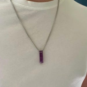 May include: A silver-toned metal chain necklace featuring a rectangular, deep purple amethyst pendant. The pendant has a smooth, polished finish and is suspended from a simple, durable link chain that rests against a plain white t-shirt.