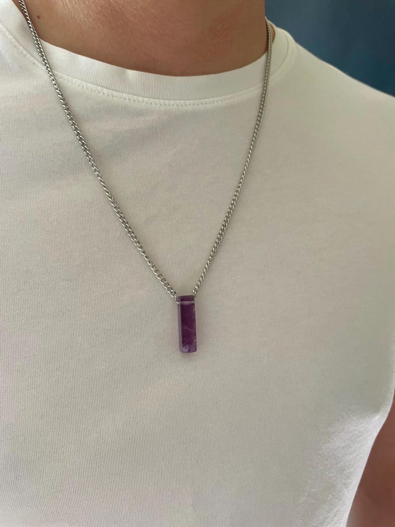 May include: A silver-toned metal chain necklace featuring a rectangular, deep purple amethyst pendant. The pendant has a smooth, polished finish and is suspended from a simple, durable link chain that rests against a plain white t-shirt.