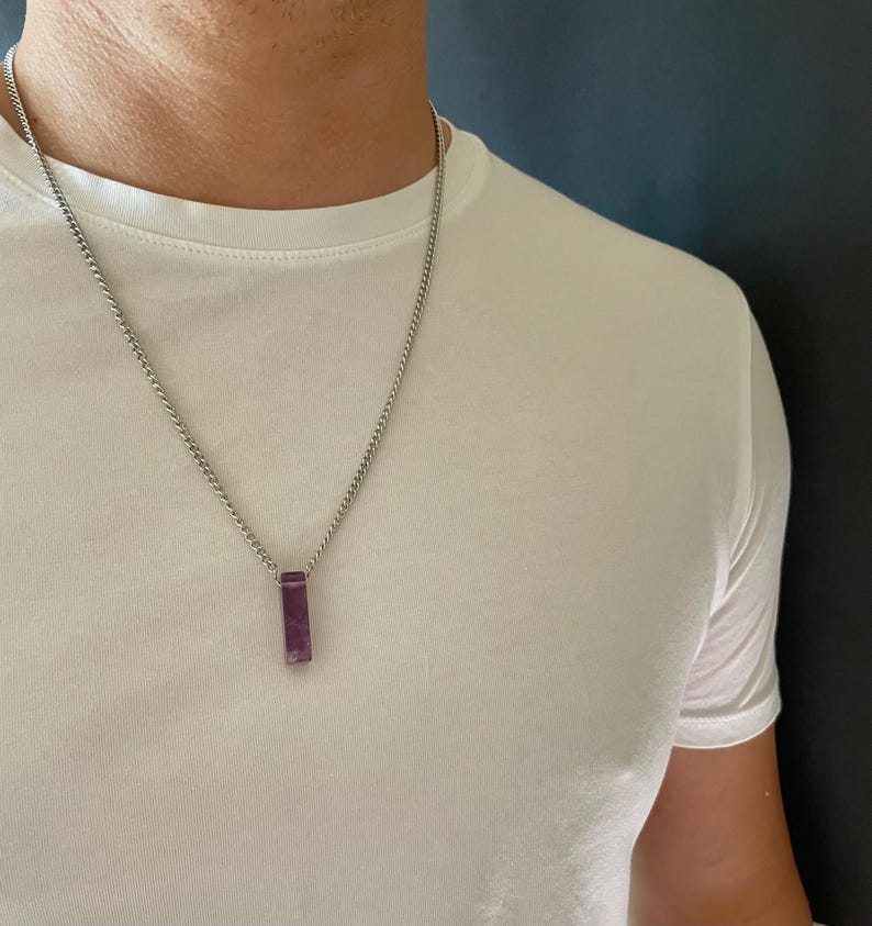May include: A silver-toned metal chain necklace featuring a rectangular, deep purple stone pendant. The pendant hangs vertically from the center of the chain, which rests against a plain white t-shirt. The stone has a smooth, polished finish.