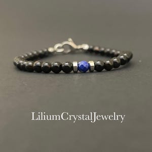 Puede incluir: Una pulsera de cuentas de ónix negro con una piedra de lapislázuli azul. La pulsera tiene un cierre plateado y cuentas plateadas. El texto "LiliumCrystalJewelry" está en la parte inferior. Una joya elegante.