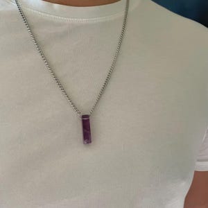 May include: A silver-toned metal chain necklace featuring a rectangular, deep purple amethyst pendant. The pendant is suspended vertically from the center of the chain, which rests against a plain white t-shirt. The stone has a smooth, polished finish.