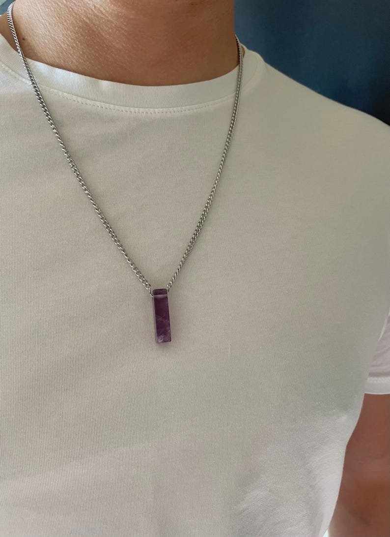 May include: A silver-toned metal chain necklace featuring a rectangular, deep purple amethyst pendant. The pendant is suspended vertically from the center of the chain, which rests against a plain white t-shirt. The stone has a smooth, polished finish.