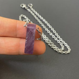 May include: A rectangular purple amethyst pendant with a polished finish, suspended from a silver-toned metal chain. The stone features natural white inclusions and is attached to the chain by a small silver-toned metal jump ring connector.