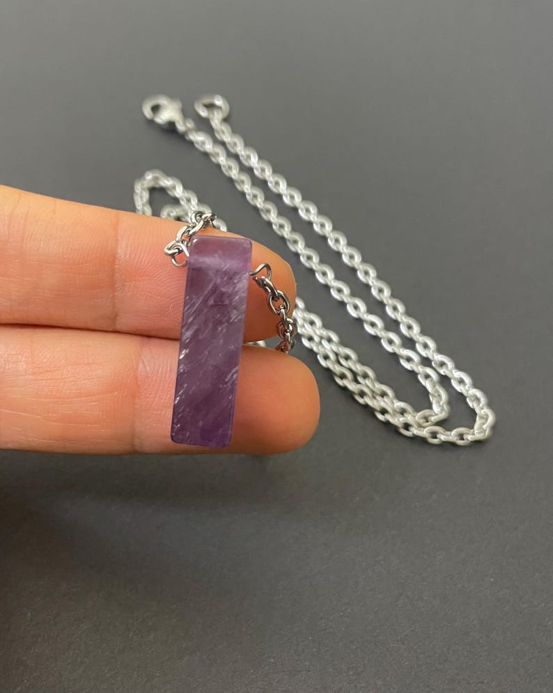 May include: A rectangular purple amethyst pendant with a polished finish, suspended from a silver-toned metal chain. The stone features natural white inclusions and is attached to the chain by a small silver-toned metal jump ring connector.
