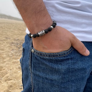 May include: A black lava rock bracelet with a silver charm. The bracelet is on a person's wrist.
