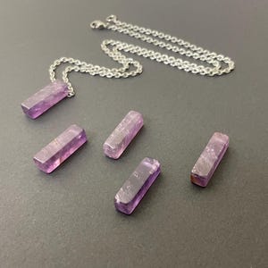 May include: A silver-toned metal chain necklace with a rectangular, translucent purple amethyst pendant, accompanied by four additional loose, matching rectangular amethyst pieces arranged in a scattered formation on a flat surface.