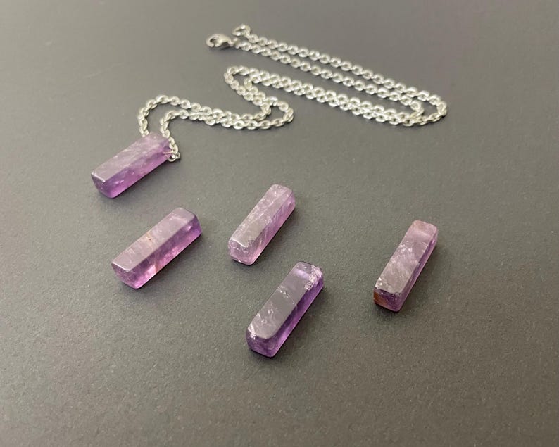 May include: A silver-toned metal chain necklace with a rectangular, translucent purple amethyst pendant, accompanied by four additional loose, matching rectangular amethyst pieces arranged in a scattered formation on a flat surface.