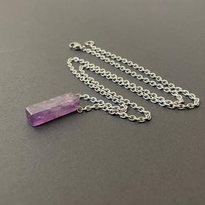 May include: A necklace featuring a rectangular, purple amethyst pendant with a rough, natural texture. The pendant is attached to a silver-toned metal chain with small, interlocking oval links and a lobster claw clasp closure at the end.