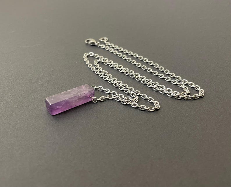 May include: A necklace featuring a rectangular, purple amethyst pendant with a rough, natural texture. The pendant is attached to a silver-toned metal chain with small, interlocking oval links and a lobster claw clasp closure at the end.